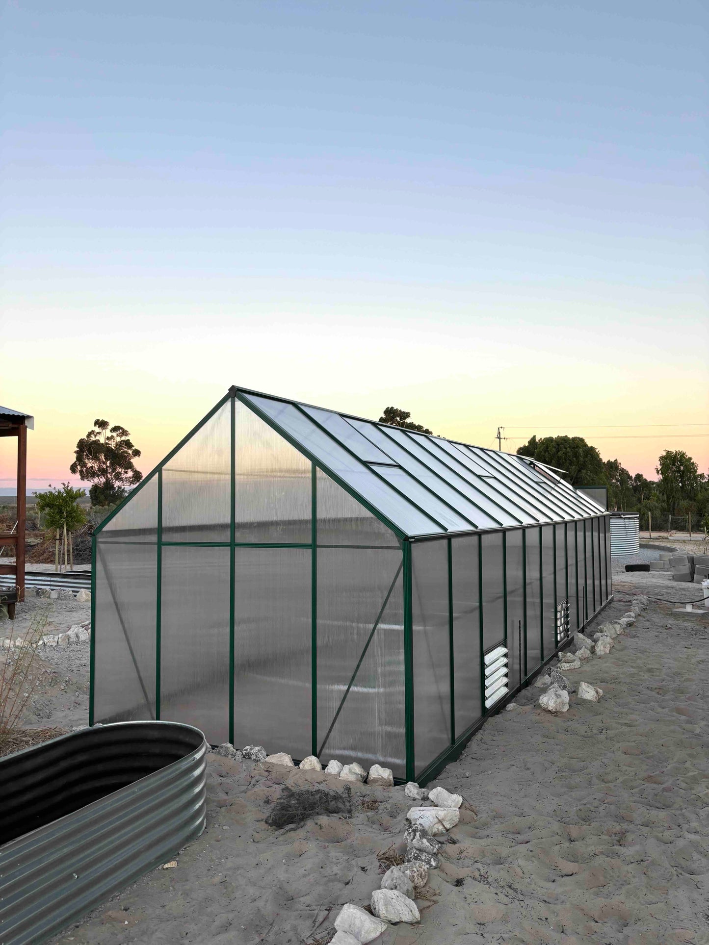 Large aluminium-frame greenhouse with polycarbonate panels in a sandy outdoor setting at sunset