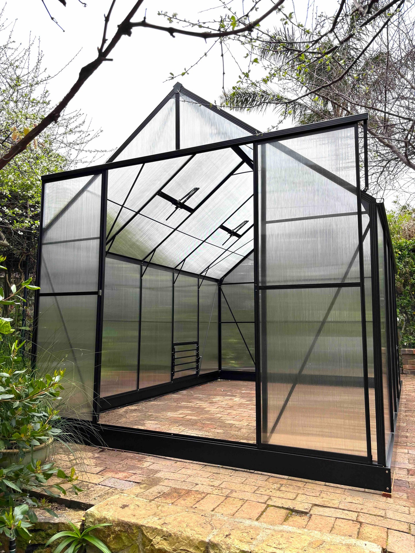 Large aluminium-frame greenhouse with clear polycarbonate panels in a garden setting