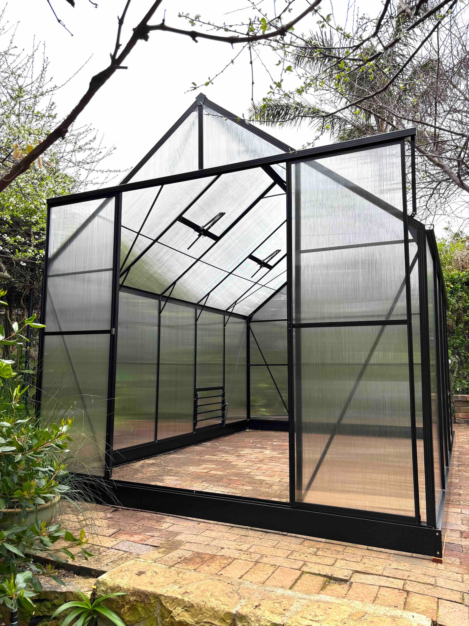 Large aluminium-frame greenhouse with clear polycarbonate panels in a garden setting