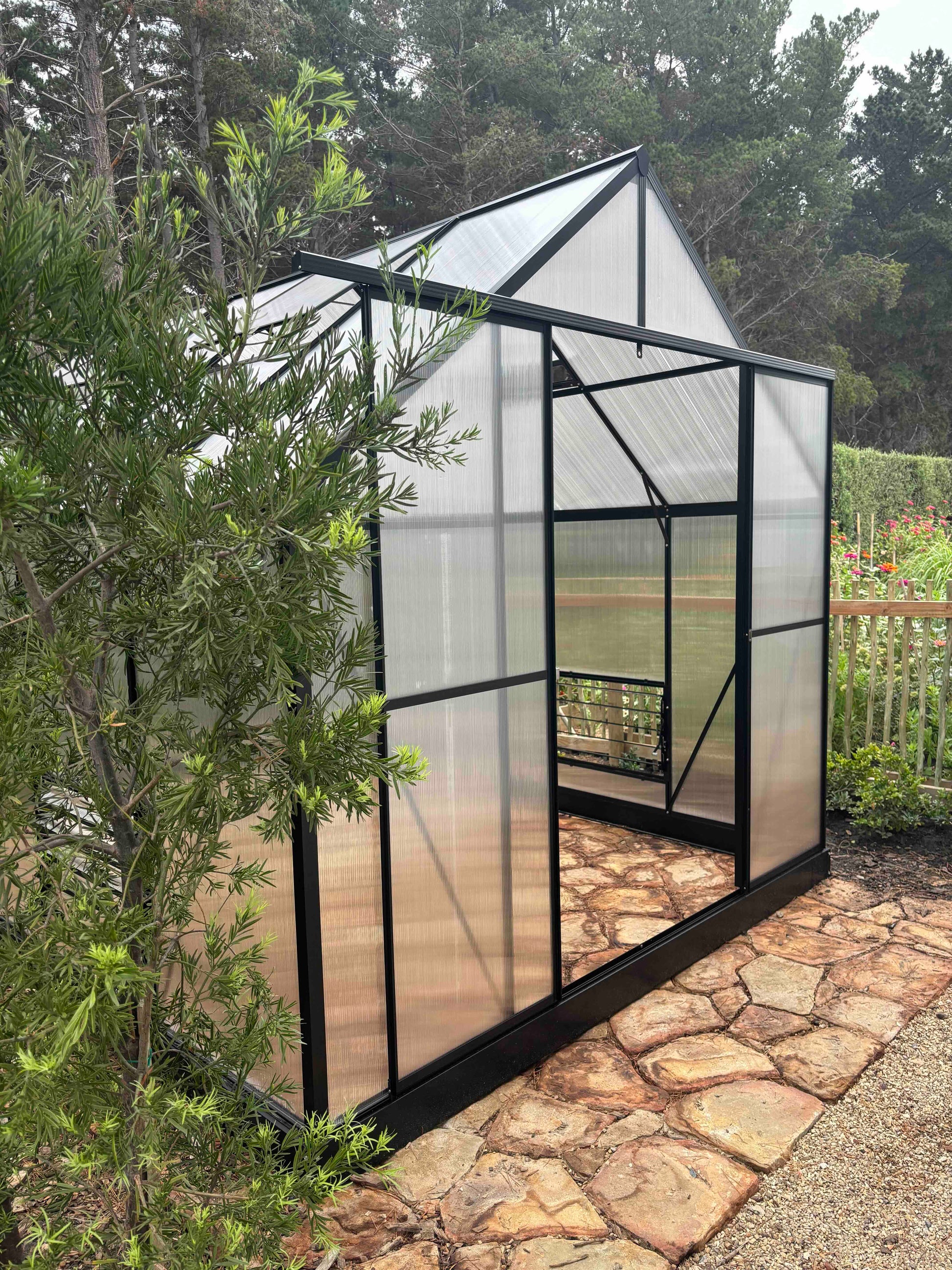 Aluminium-frame greenhouse with polycarbonate panels on stone path in garden setting